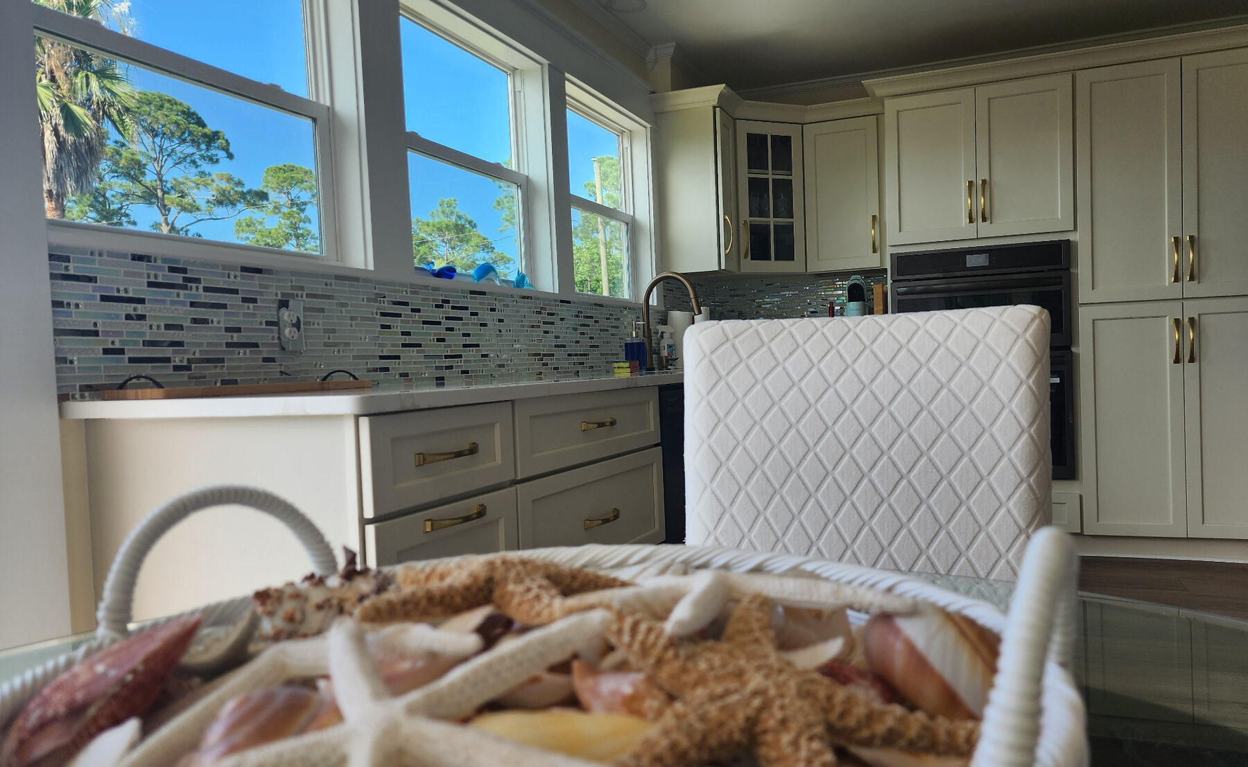 Kitchen counters with tile backsplash, gold fixtures, and a basket of seashells and starfish on the dining table in the foreground